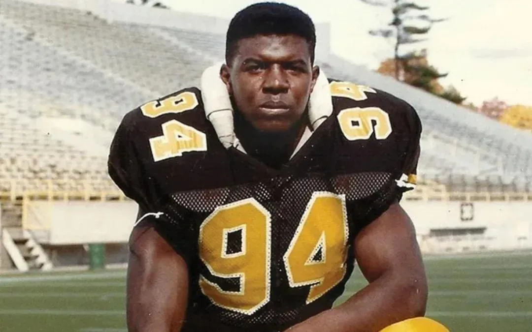 A vintage photo shows a Black American football player in a black and gold uniform with the number 94 on his chest and…