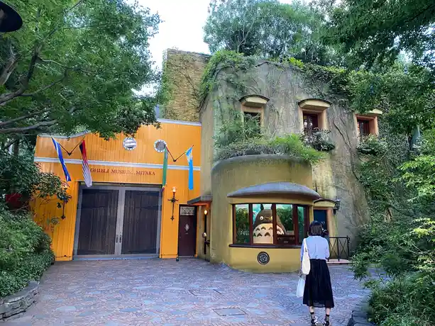 A person stands on a cobblestone path facing the Ghibli Museum in Mitaka, Japan, where a large, friendly Totoro plush figure…