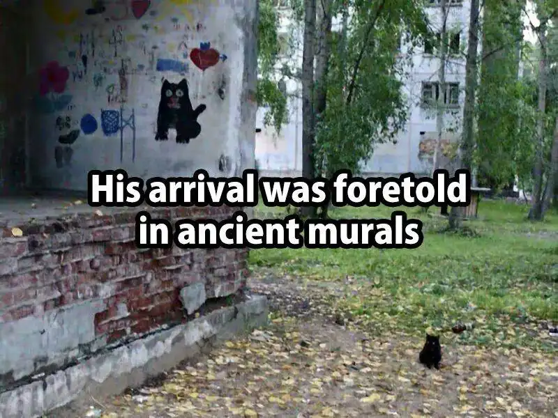A small black cat sits on a leafy path near a brick wall, with a faded mural of a black cat painted on the wall behind it…