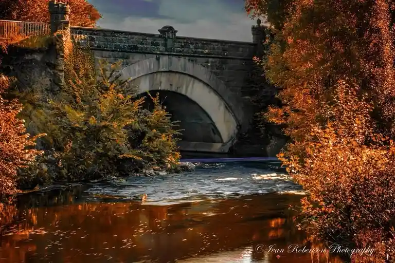 A scenic autumn photograph of a stone arch bridge over a river, framed by vibrant orange foliage on both banks. The water…