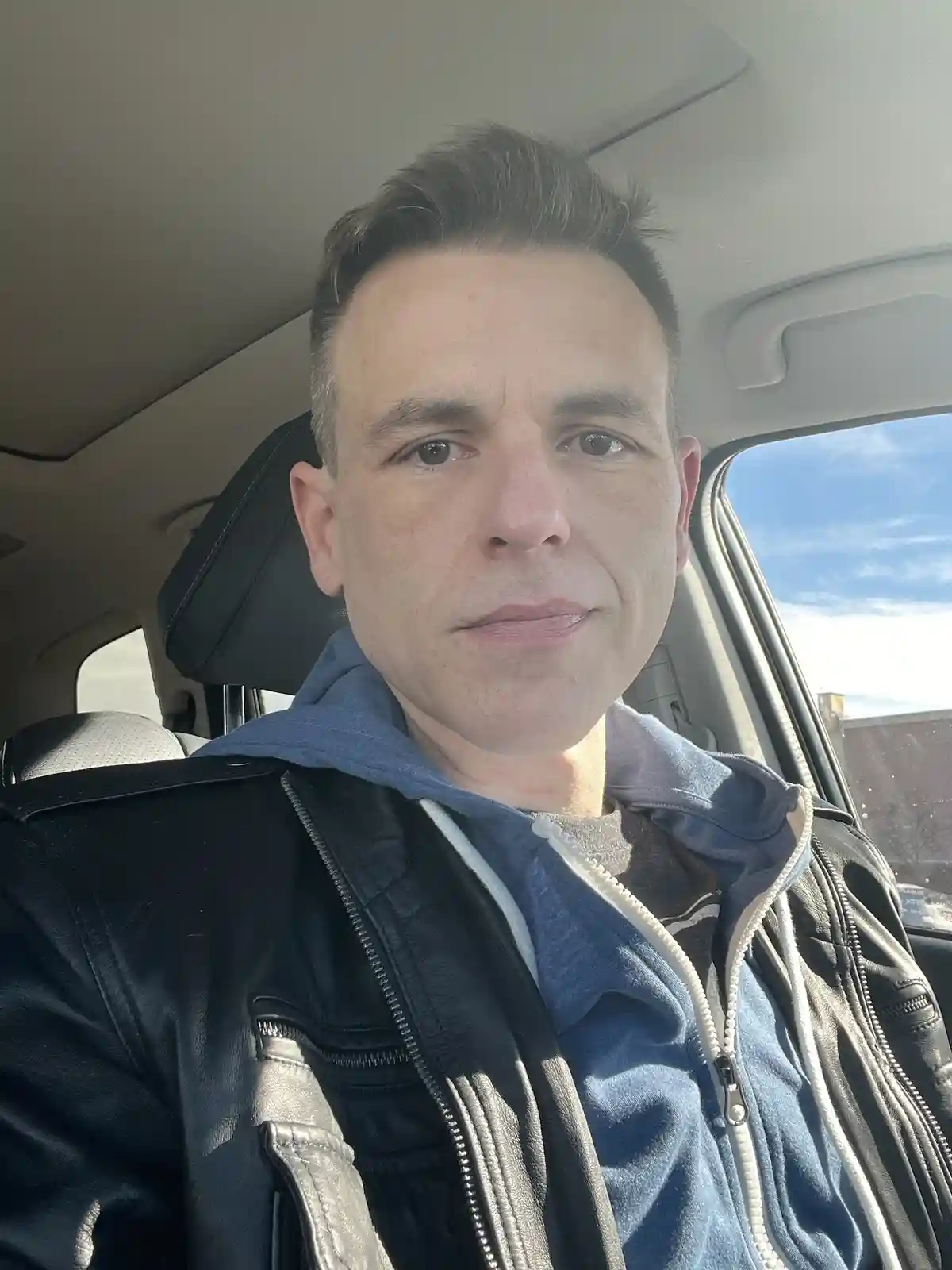 A man with short dark hair sits in the driver's seat of a car, wearing a black leather jacket over a blue hoodie. He looks…