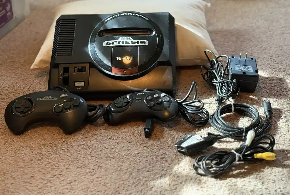 A black Sega Genesis console with two controllers, power adapter, and cables laid out on a carpeted floor, with the text…