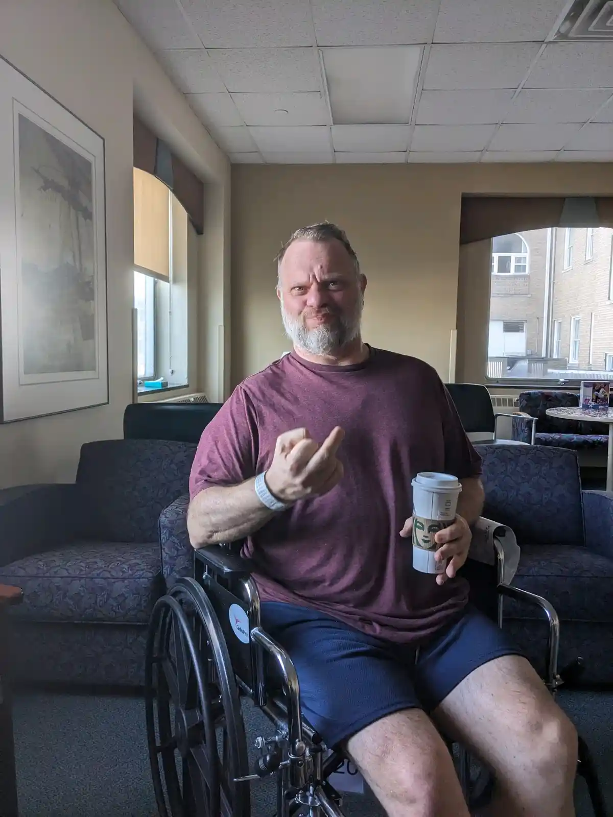 A middle-aged man with a beard sits in a wheelchair in a waiting room, holding a Starbucks coffee cup and making a defiant…