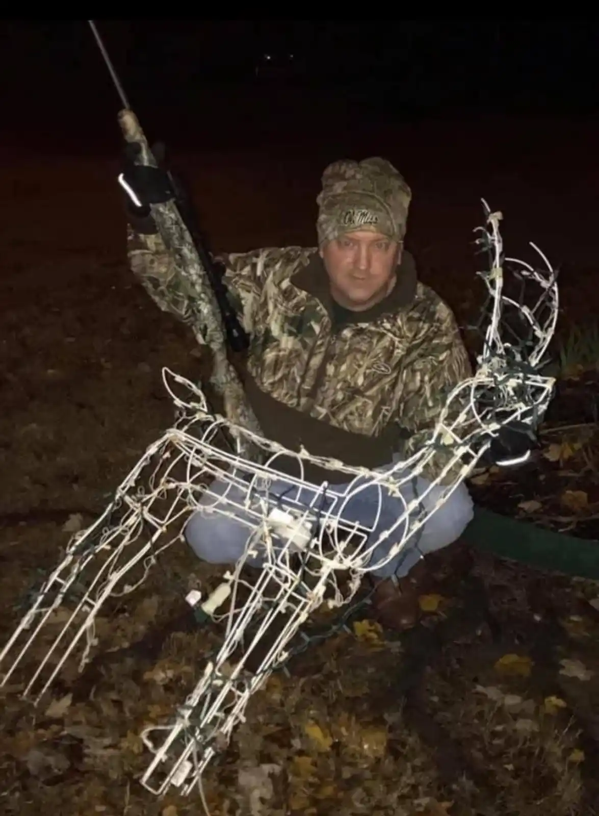 A man in camouflage clothing and a beanie kneels outdoors at night holding a rifle, posing beside a large, wireframe…