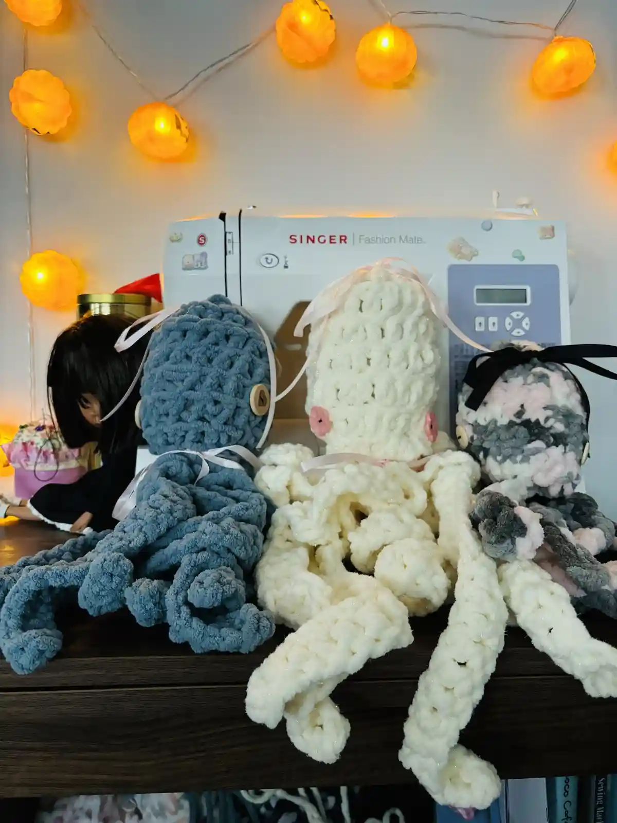 Three crocheted octopus plushies in blue, white, and gray/pink sit on a wooden surface in front of a white Singer sewing…