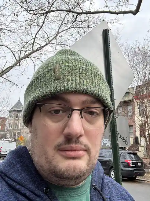 A man with glasses and a green knit beanie stands outdoors next to a blank, back-facing traffic sign. He has a neutral…