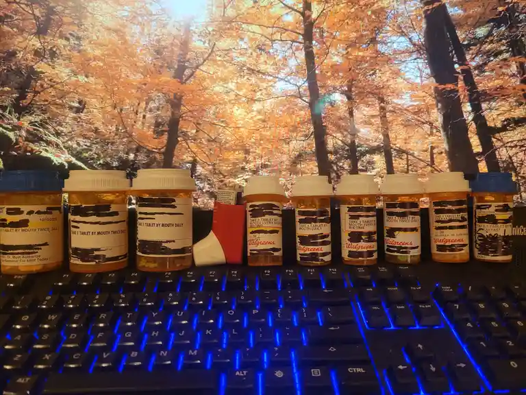 Multiple prescription pill bottles with obscured labels are lined up on a blue-lit computer keyboard, with a bright autumn…