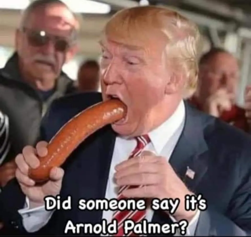 A man in a suit and tie is shown eating a large sausage with his mouth wide open. Behind him, another man wearing sunglasses…