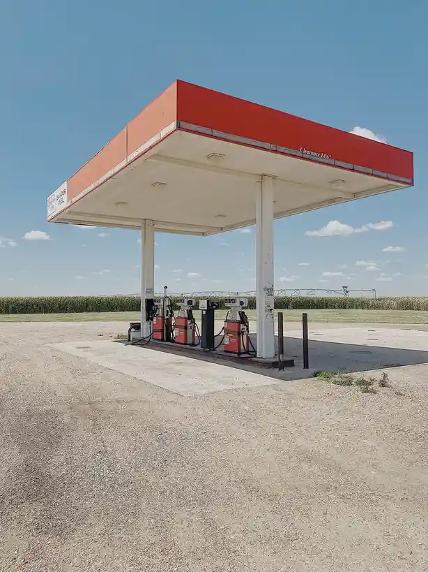 A lone, weathered gas station with red and white canopy stands in a vast, open field under a clear blue sky, surrounded by…