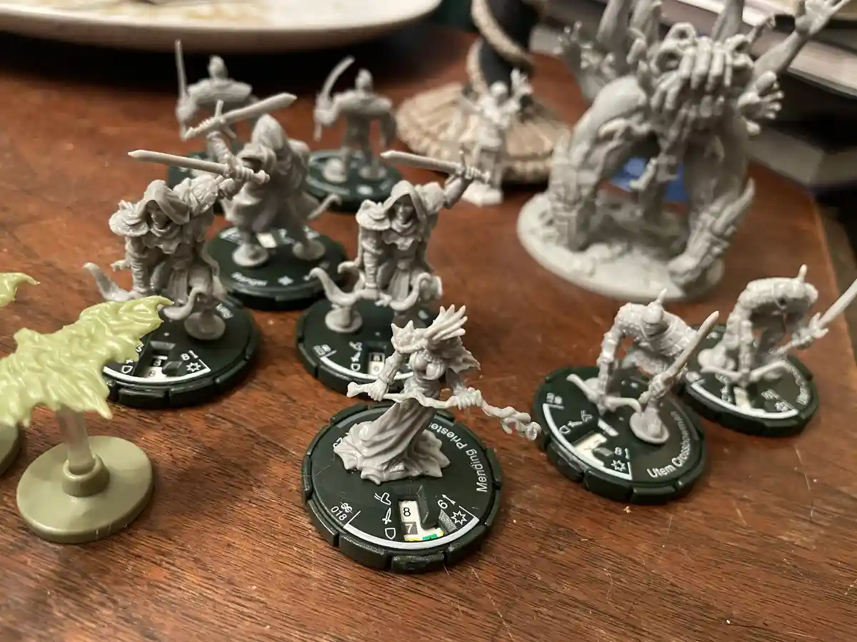 A collection of unpainted fantasy miniatures on circular green bases are arranged on a wooden table, with one central figure…
