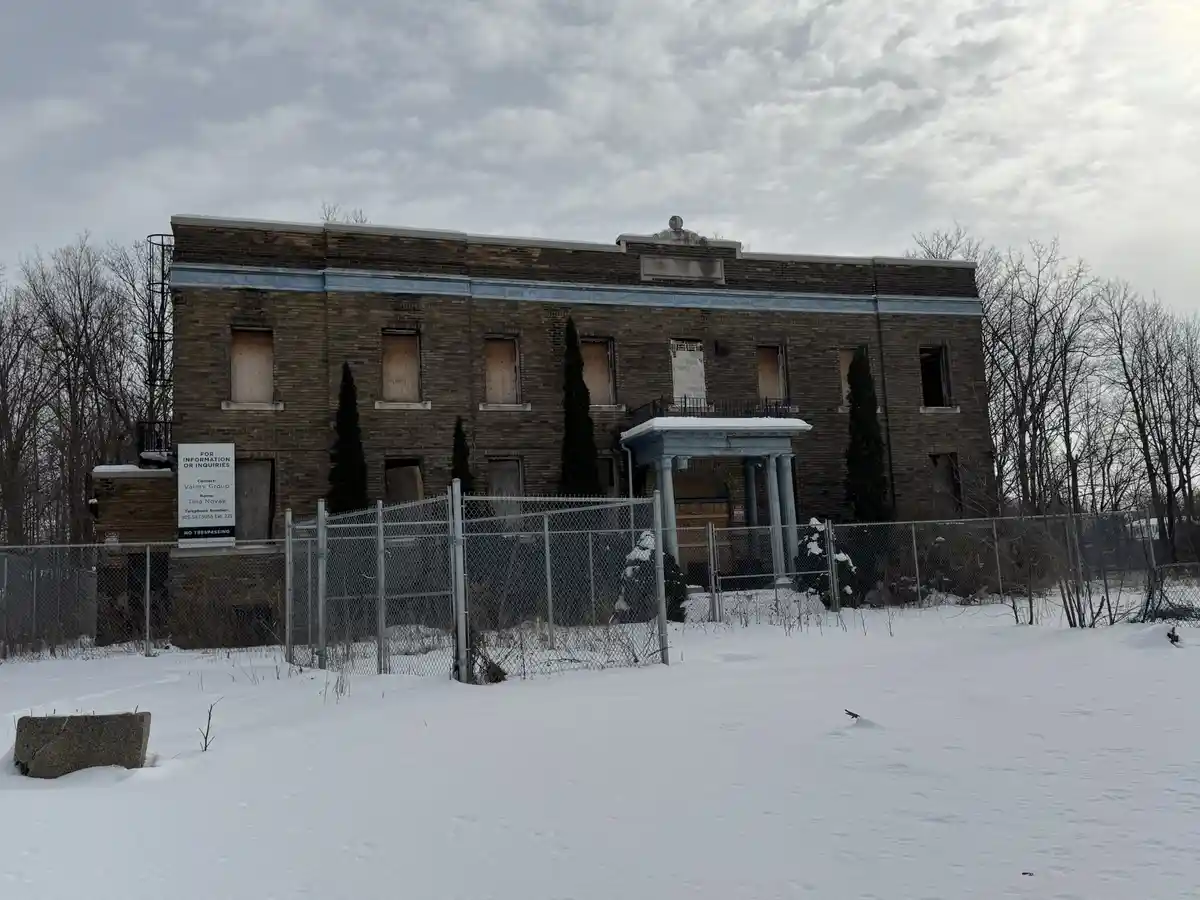 A dilapidated, snow-covered brick building with boarded-up windows and a chain-link fence in front. A real estate sign on…