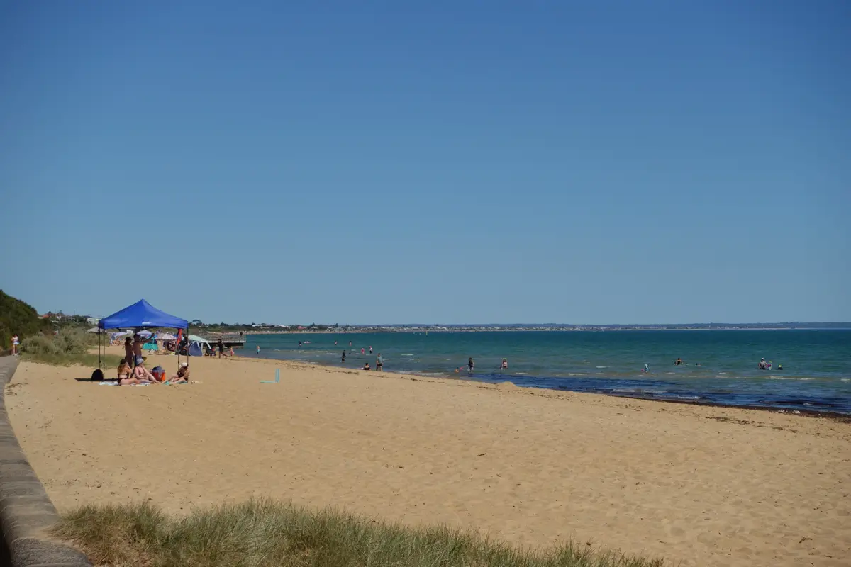 A wide beach scene under a clear blue sky with people sunbathing under a blue pop-up tent and swimming in the ocean. The…