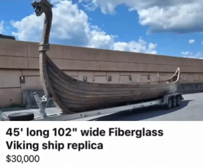 A large fiberglass Viking ship replica with a dragon head at the prow is parked on a multi-axle trailer in front of a beige…