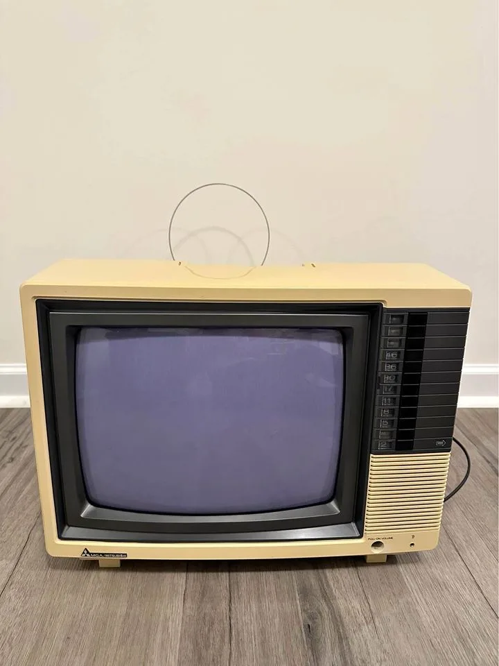 A beige, vintage-style CRT television with a blank purple screen sits on a wooden floor against a plain wall. The TV has a…