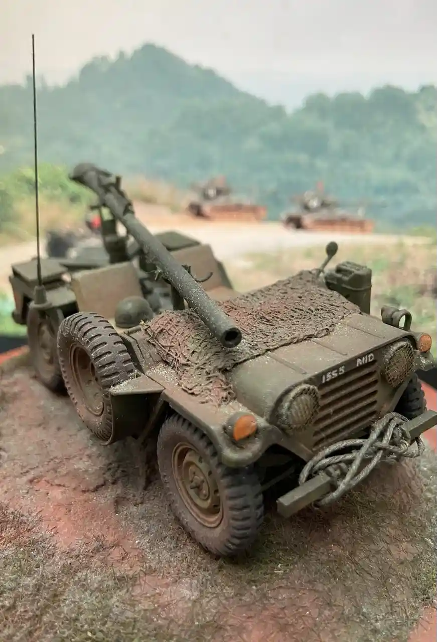 A detailed scale model of a military jeep mounted with a large gun, covered in camouflage netting and dirt, sits on a…