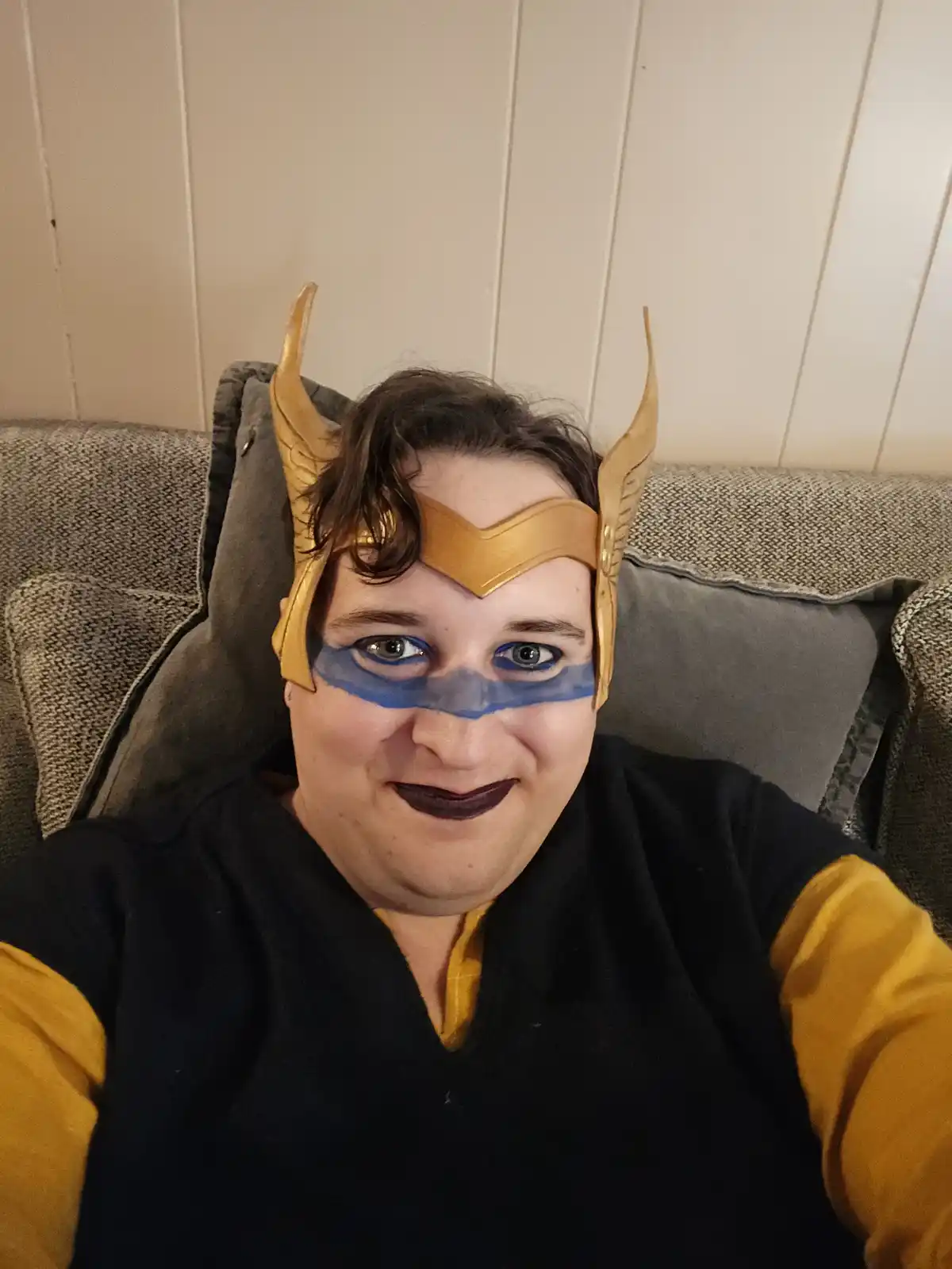 A person sits on a gray couch wearing a golden Thor-style helmet with horns and blue eye paint, smiling while dressed in a…