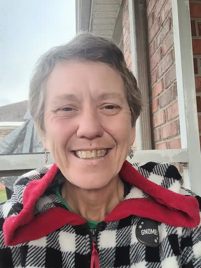 A smiling older woman with short gray hair wears a black-and-white checkered fleece jacket with a red collar and a black…