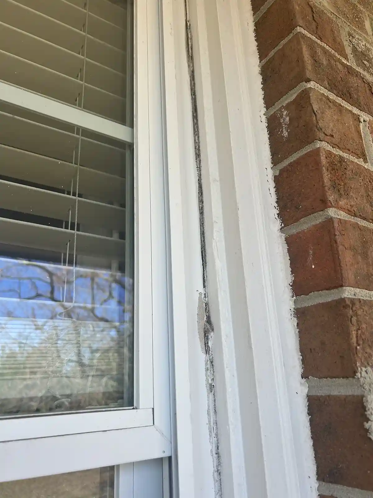 A close-up photo shows a white window frame set into a red brick wall, with visible cracks and gaps filled with messy…