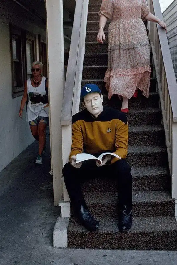 A man in a Star Trek uniform and a blue LA baseball cap sits on outdoor stairs reading a book with a blank, focused…