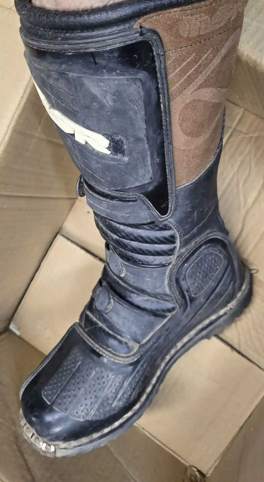 A close-up photo shows a heavily worn, dirty black and brown motorcycle boot resting on cardboard. The boot has visible…