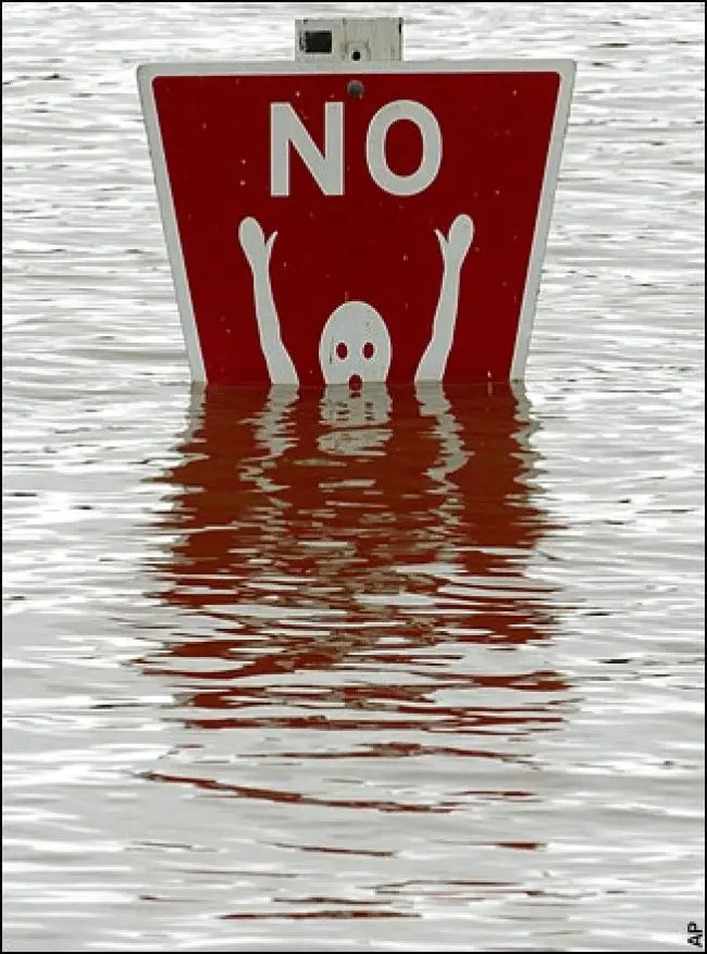 A red rectangular traffic sign with the word 'NO' in white letters is partially submerged in floodwater, with a white…