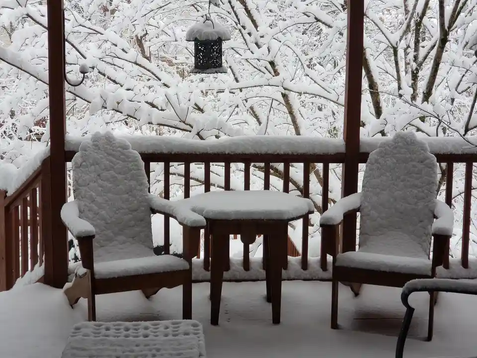 A snow-covered wooden deck features two chairs and a table, all blanketed in fresh snow, with snow-laden trees visible in…