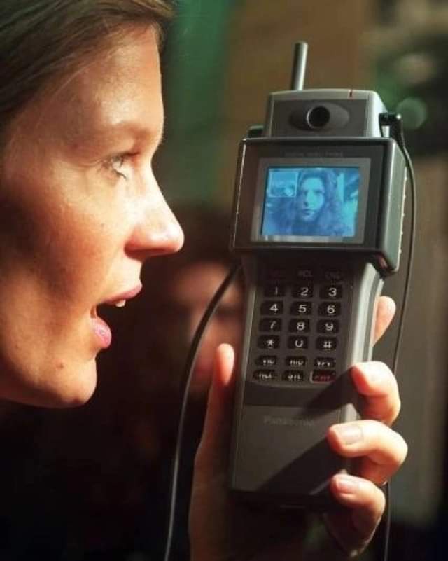 a woman holds an old, bulky Panasonic phone with a small screen showing a man's face, looking shocked as she stares at the…