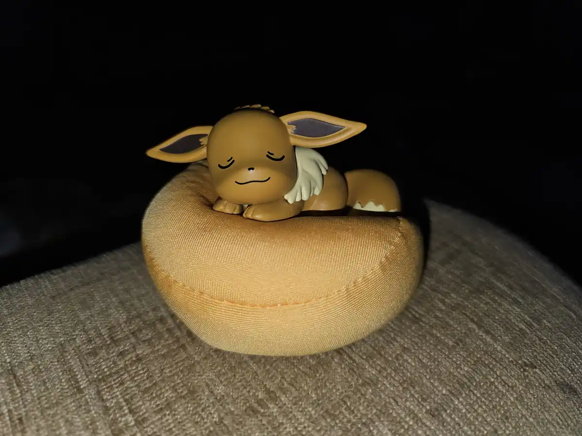 A small, tan Eevee figurine with closed eyes and a serene expression is lying on a tan, fabric-covered, crescent-shaped…