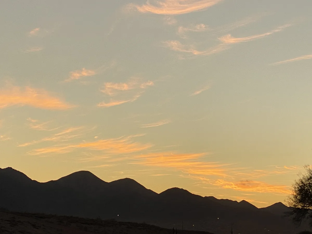 A serene landscape photo shows a mountain range silhouetted against a sky painted with soft orange and yellow hues from a…