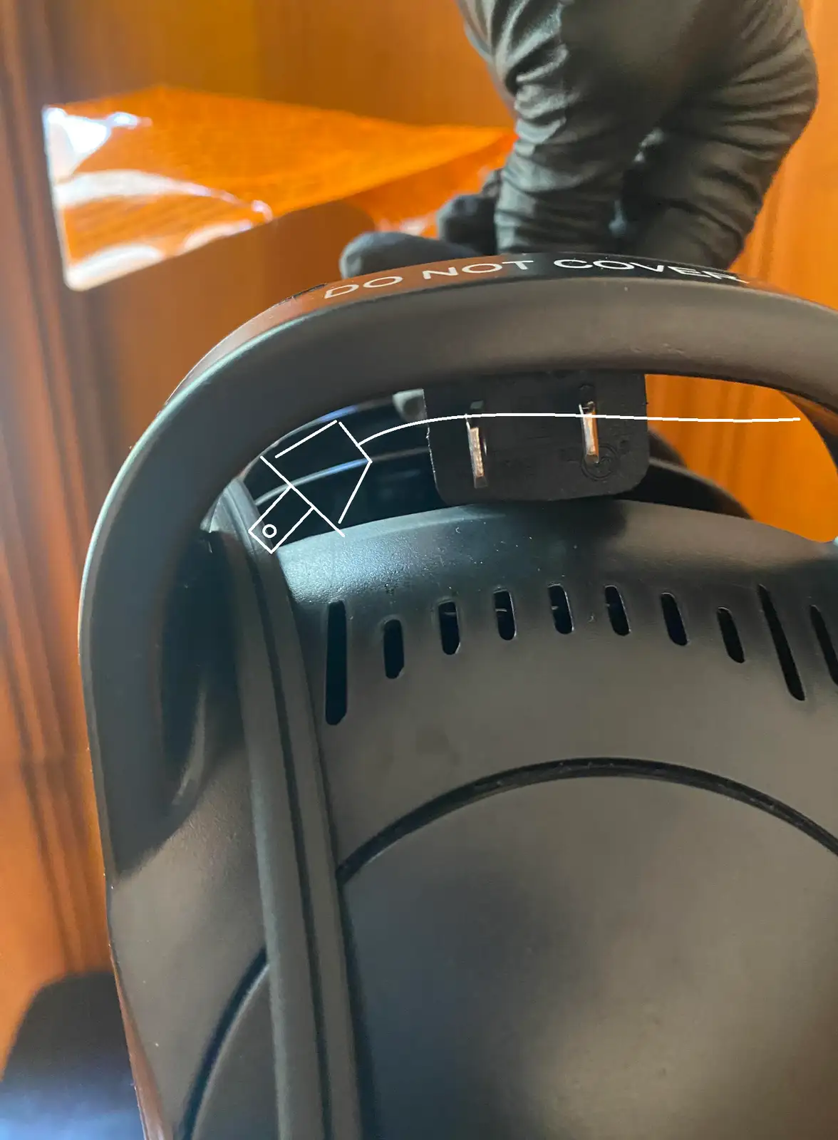 A close-up photo shows a black appliance with a power plug inserted into its top slot. A white line drawn on the image…