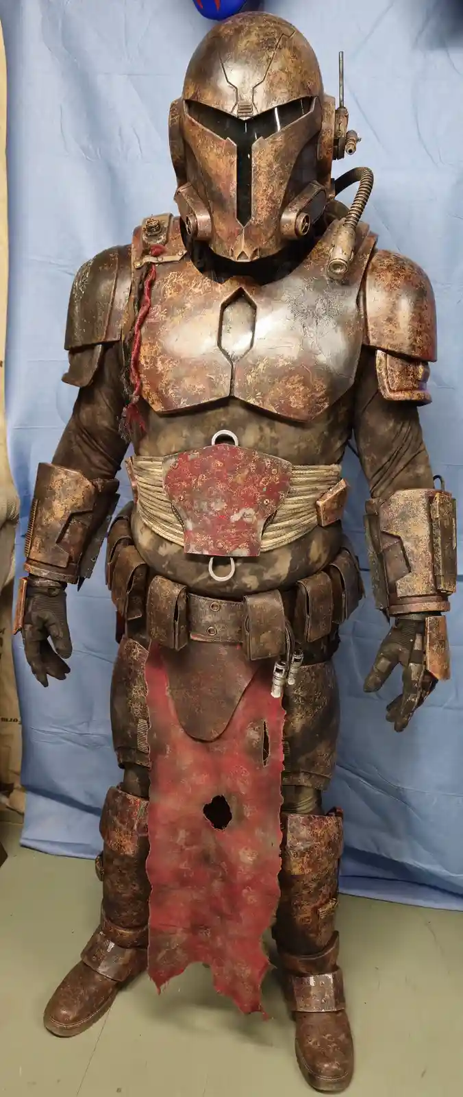 A full-body, heavily weathered and rusted Mandalorian-style armor suit stands against a blue backdrop, featuring a helmet…