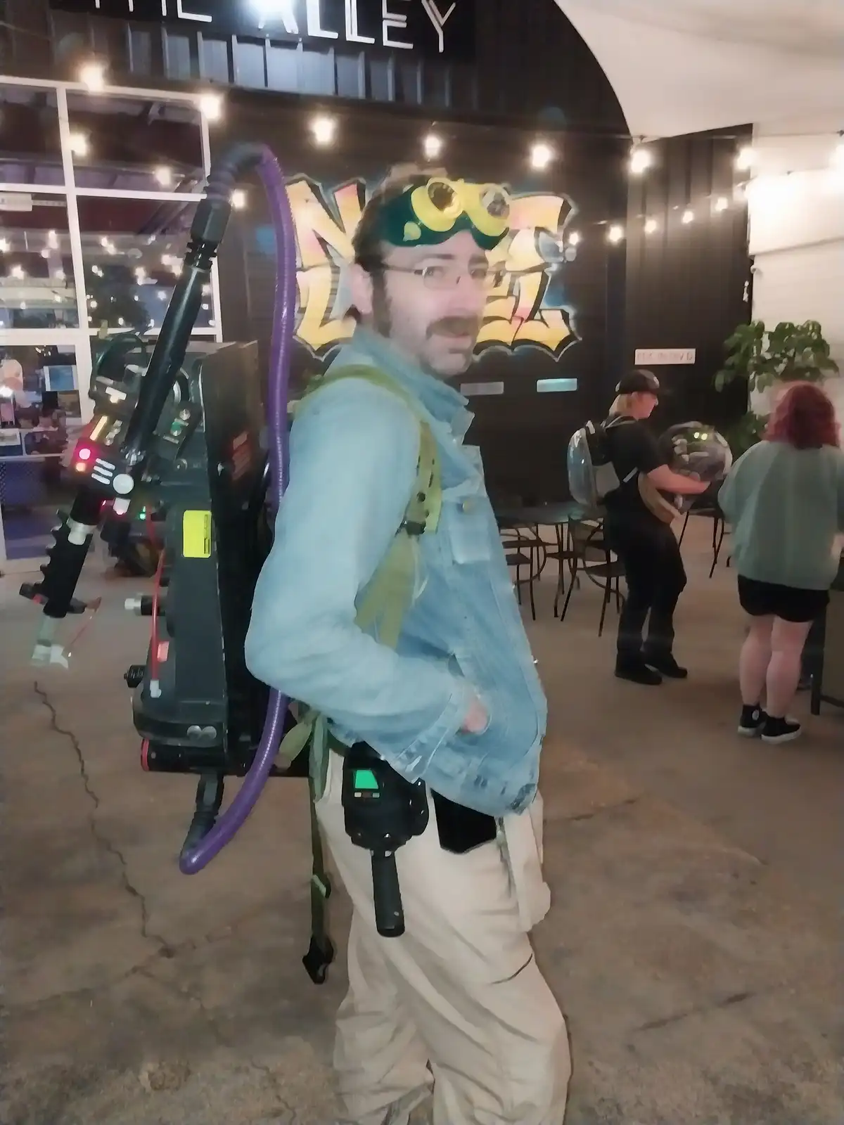 A man in a denim jacket and beige pants wears yellow goggles and carries a large, purple-hosed ghost-busting device on his…