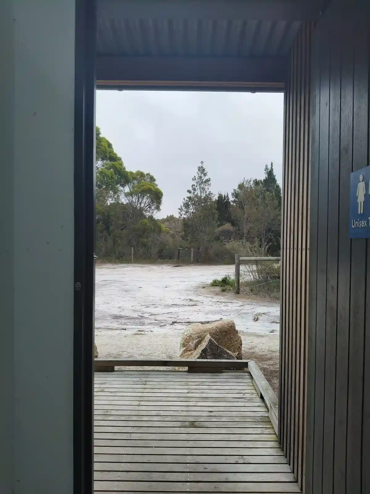 A view from inside a unisex restroom looking out through an open doorway onto a wooden walkway leading to a gravel area with…