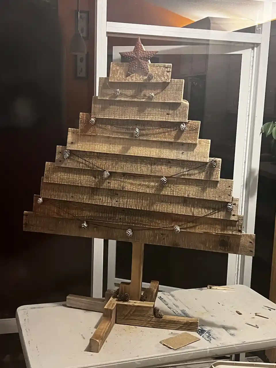 A handmade, rustic wooden Christmas tree made from stacked weathered planks, decorated with pinecones strung on wire and…
