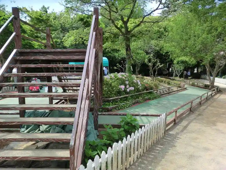 An outdoor miniature train track with a wooden staircase leading up to it, surrounded by greenery and pink flowers. The…