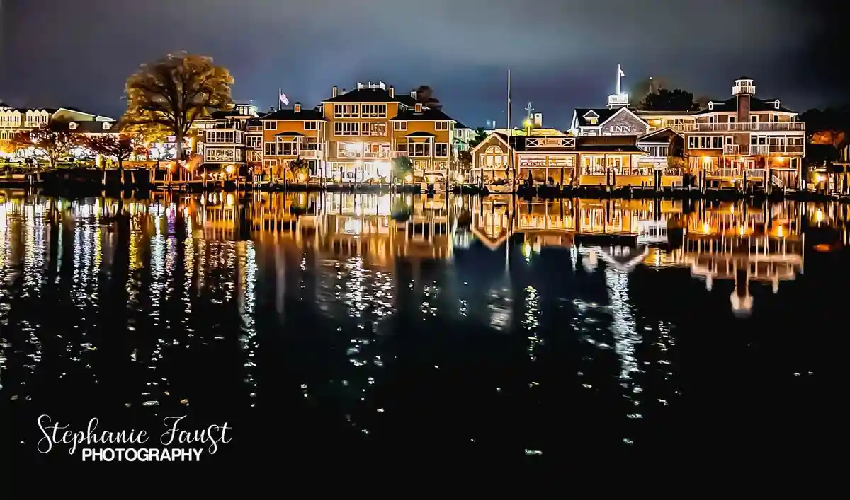 A nighttime photograph of waterfront buildings with warm lights reflecting on dark water, featuring a watermark in the…