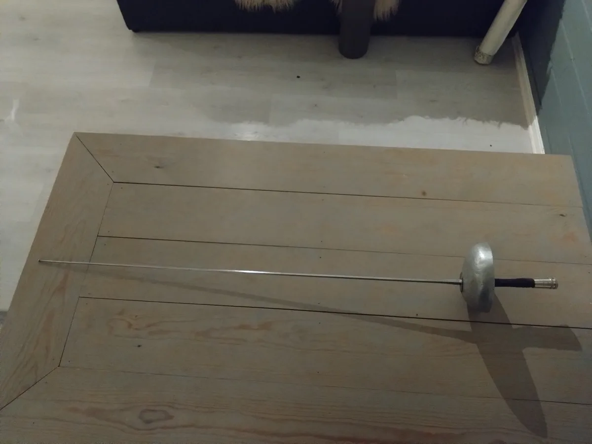 A fencing sword with a metallic blade and black grip lies horizontally on a light-colored wooden table. In the background, a…