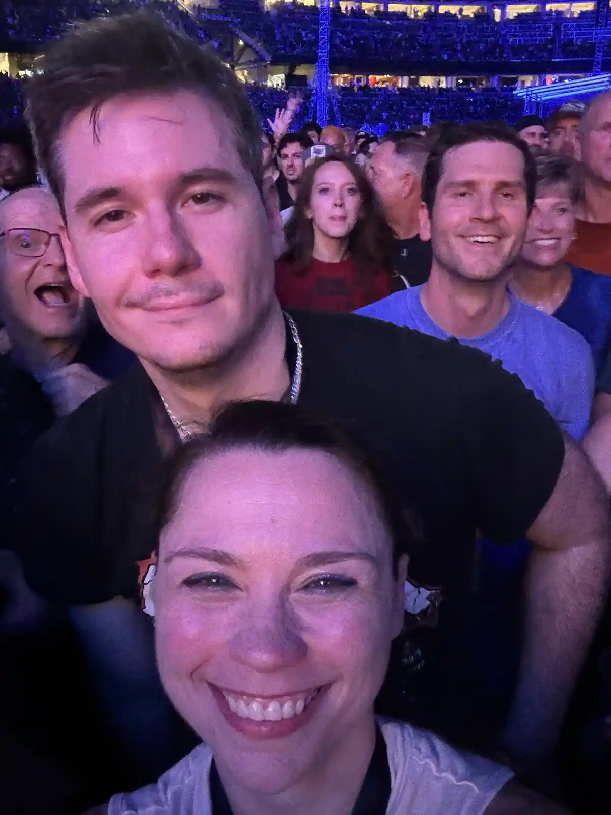 A selfie taken at a crowded concert or arena shows a smiling woman in the foreground and a man behind her, both looking at…