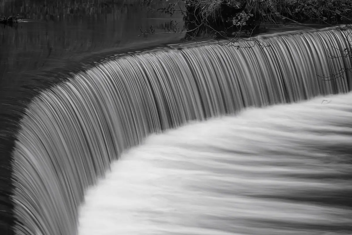 A black-and-white photograph of a waterfall with motion blur, showing water cascading over a curved ledge into a frothy pool…