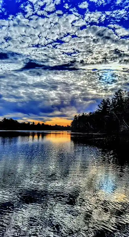 A highly saturated, vertically oriented photograph of a lake at sunset, showing dramatic blue and white clouds reflected in…