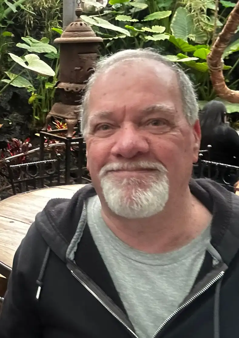 A middle-aged man with a white goatee and gray hair smiles at the camera while seated at a wooden table in a lush, green…