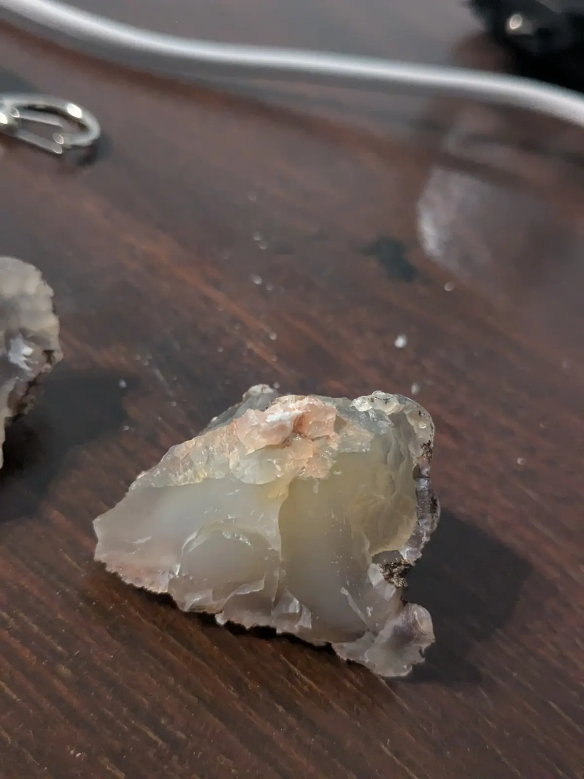 A close-up photo shows a rough, translucent, light-colored rock with some pinkish mineral inclusions resting on a dark…