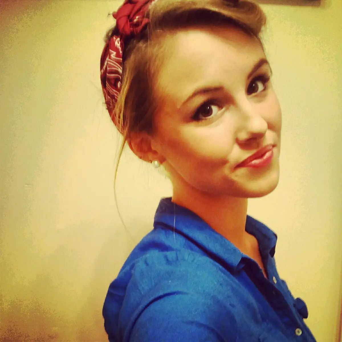 A young woman with light brown hair styled in a vintage updo with a red patterned headscarf, wearing a blue collared shirt…