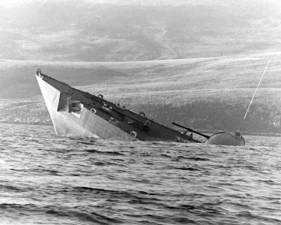 A black-and-white photo shows a ship heavily tilted and sinking in water, with its bow high in the air and stern submerged…
