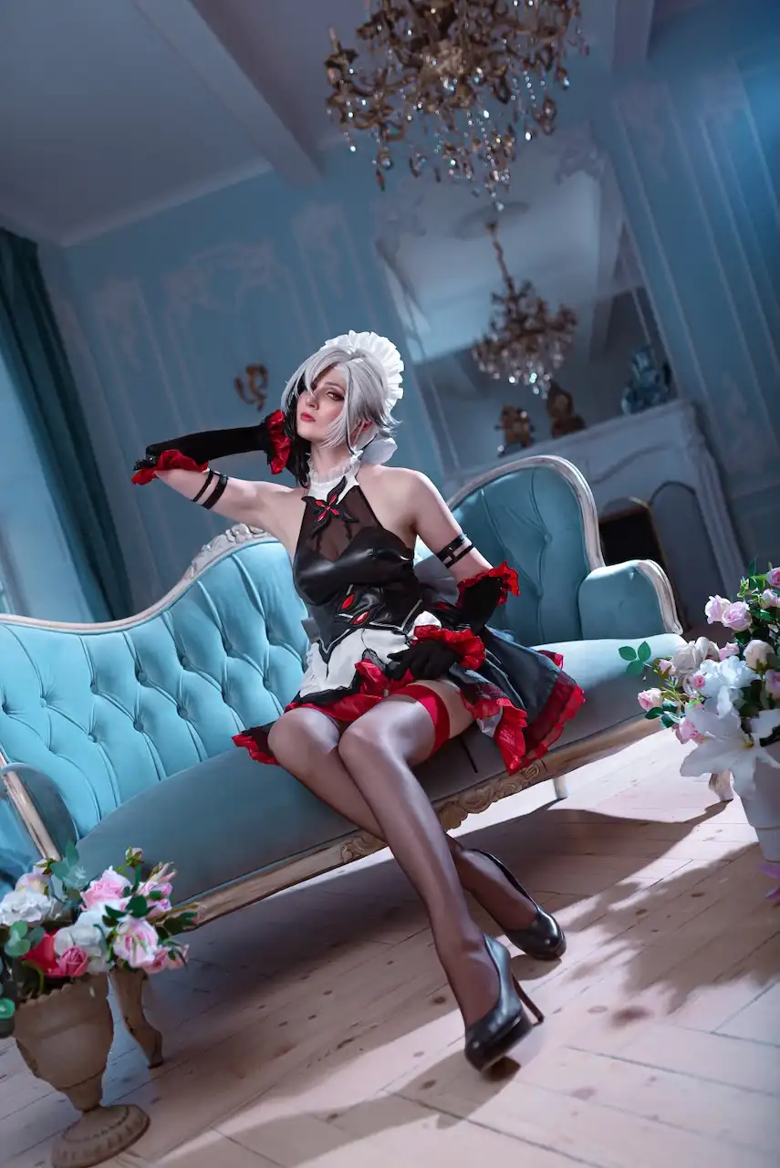 A person in a maid outfit with white hair and red accents poses on a blue velvet couch in a luxurious, ornate room with…
