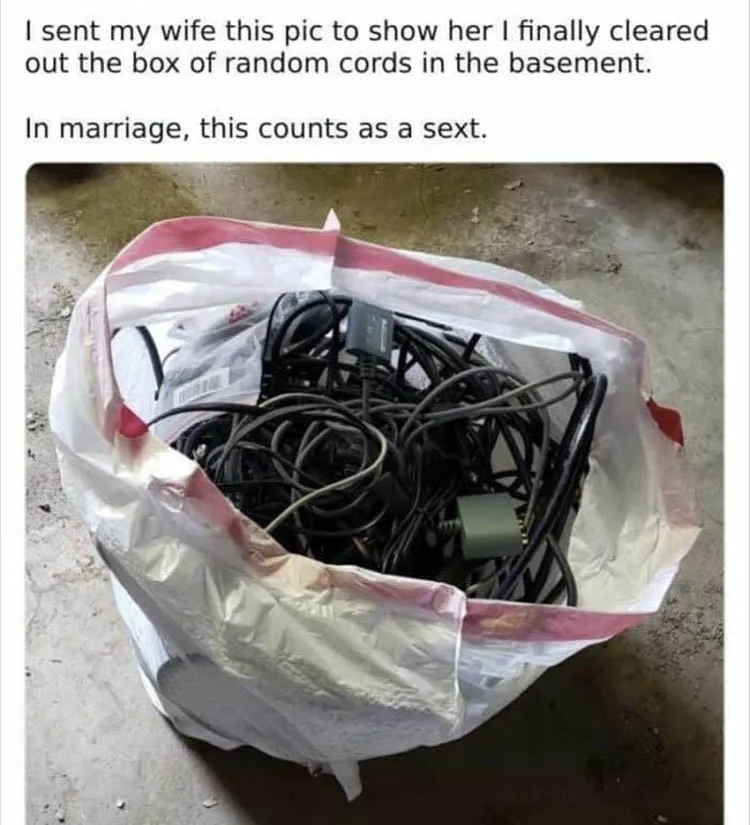 A large white plastic bag filled with a tangled mess of black and gray electrical cords and adapters sits on a concrete…