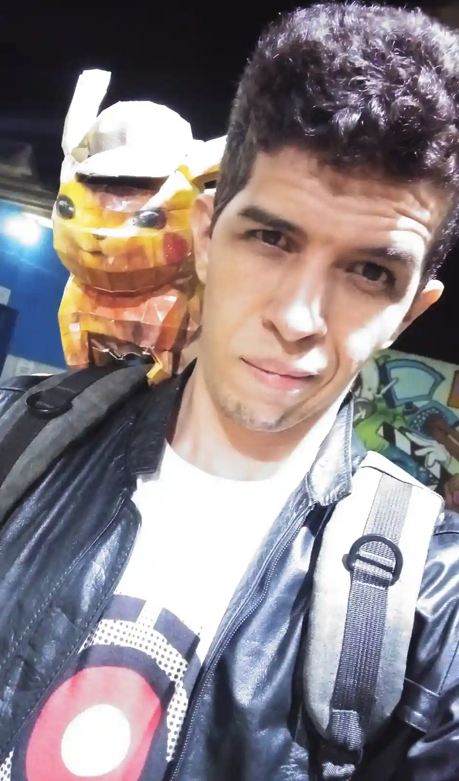 A man in a black leather jacket and white t-shirt takes a selfie, looking directly at the camera with a skeptical…