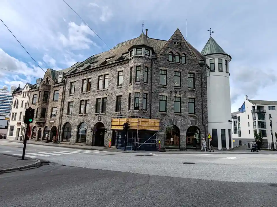 A wide-angle street photo shows a large, historic stone building with arched ground-floor windows and a prominent white…