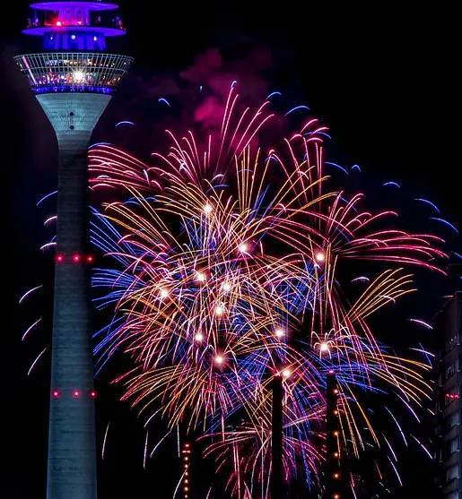 A nighttime photo of a tall, illuminated observation tower with a vibrant fireworks display exploding in front of it. The…