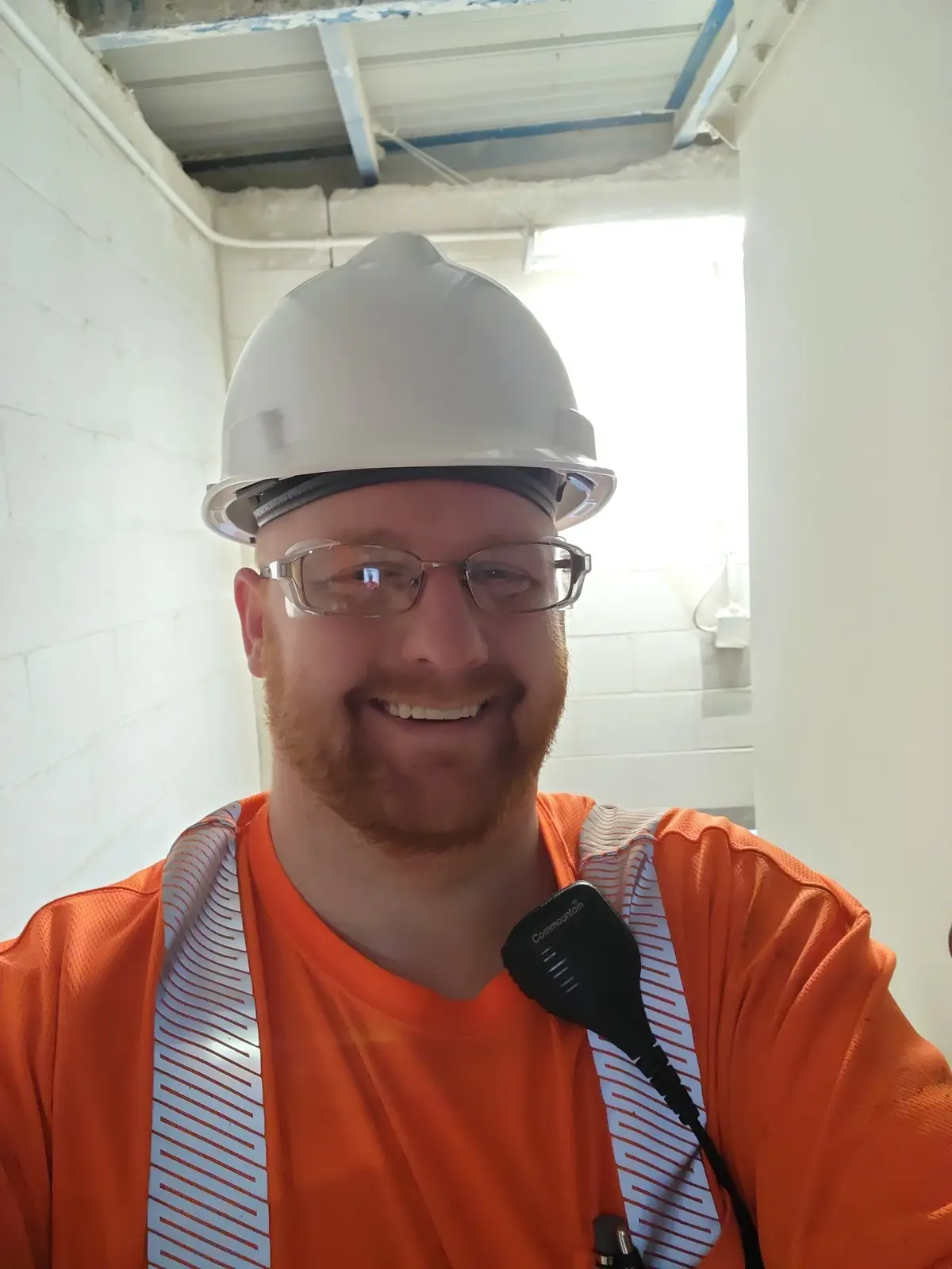 A smiling man with a red beard and glasses wears a white hard hat and an orange safety vest with a black 'Commountain' radio…